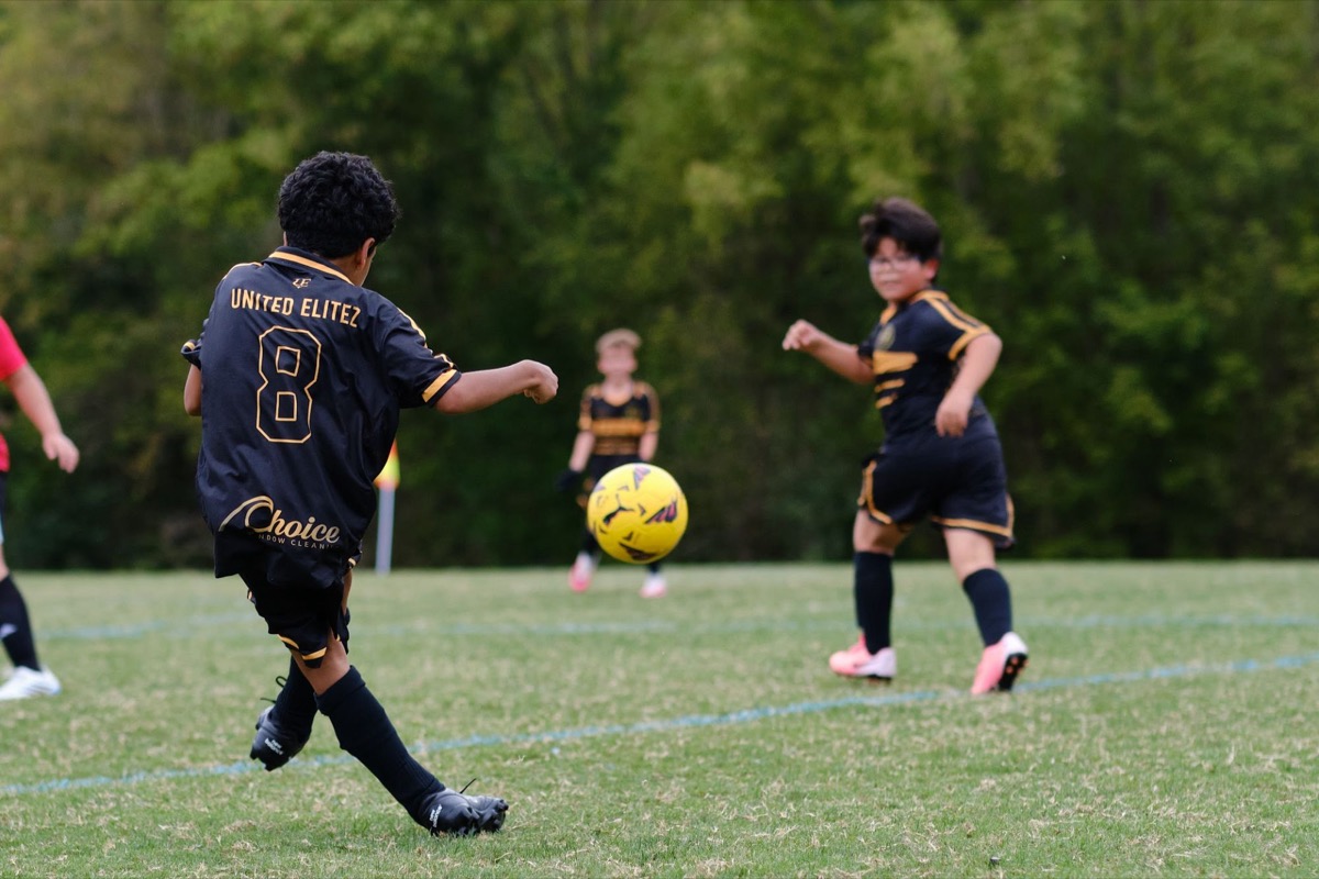 A United Elitez FC player striking the ball during a match, wearing a jersey with the Choice Window Cleaning sponsor logo